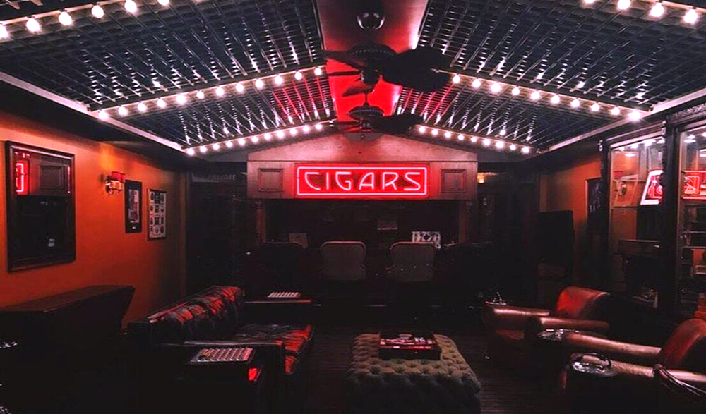 A man cave with a neon cigar sign hanging above a bar stocked with alcohol and glasses